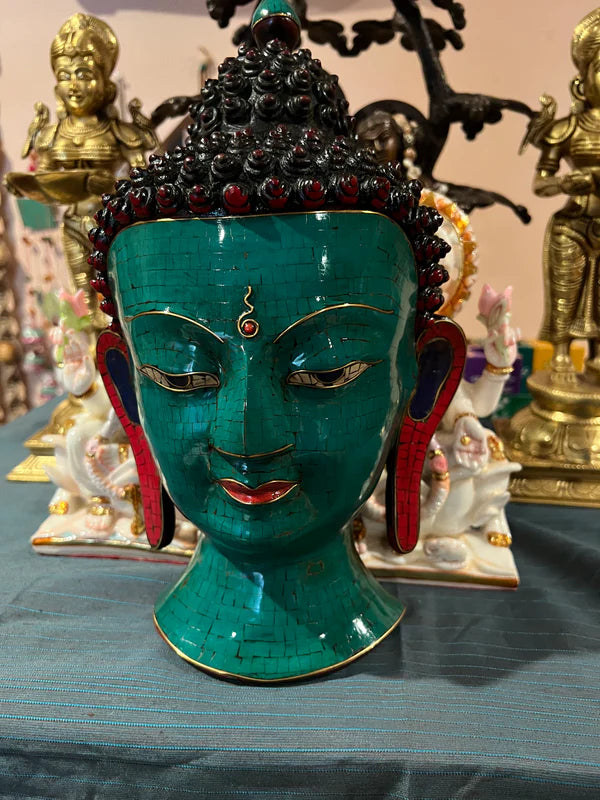 Displayed elegantly on a cloth surface is the Buddha mask green lapis from Saanskrityam Handicraft Store, featuring intricate red and black details. In the background, golden and ivory figures complement the scene's spiritual and decorative ambiance.