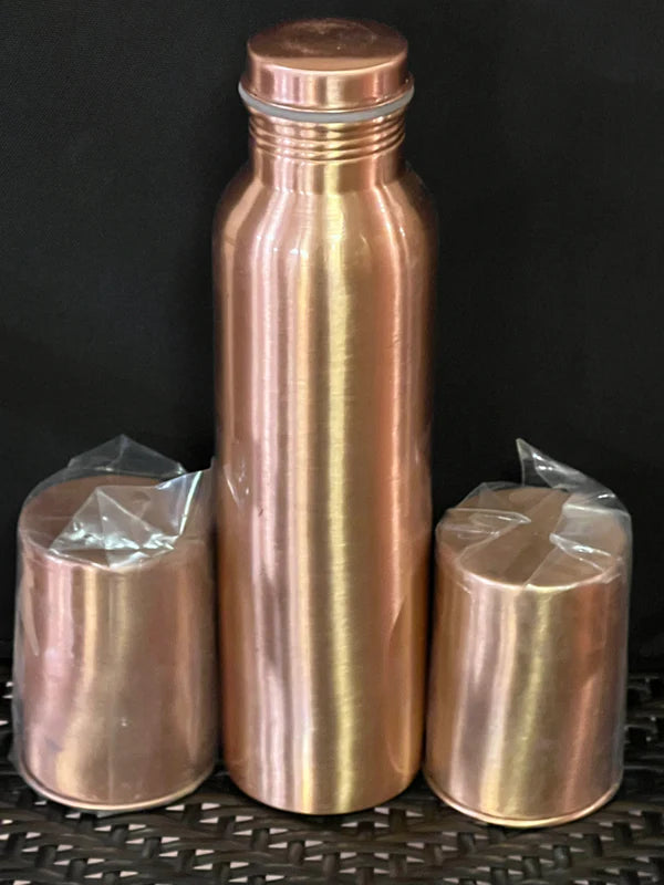 The Saanskrityam Handicraft Store presents a Copper water bottle with 2 tumblers, perfect as gifts for the holiday season. Wrapped in plastic and set on a woven mat, the arrangement features cups flanking the bottle against a dark backdrop.