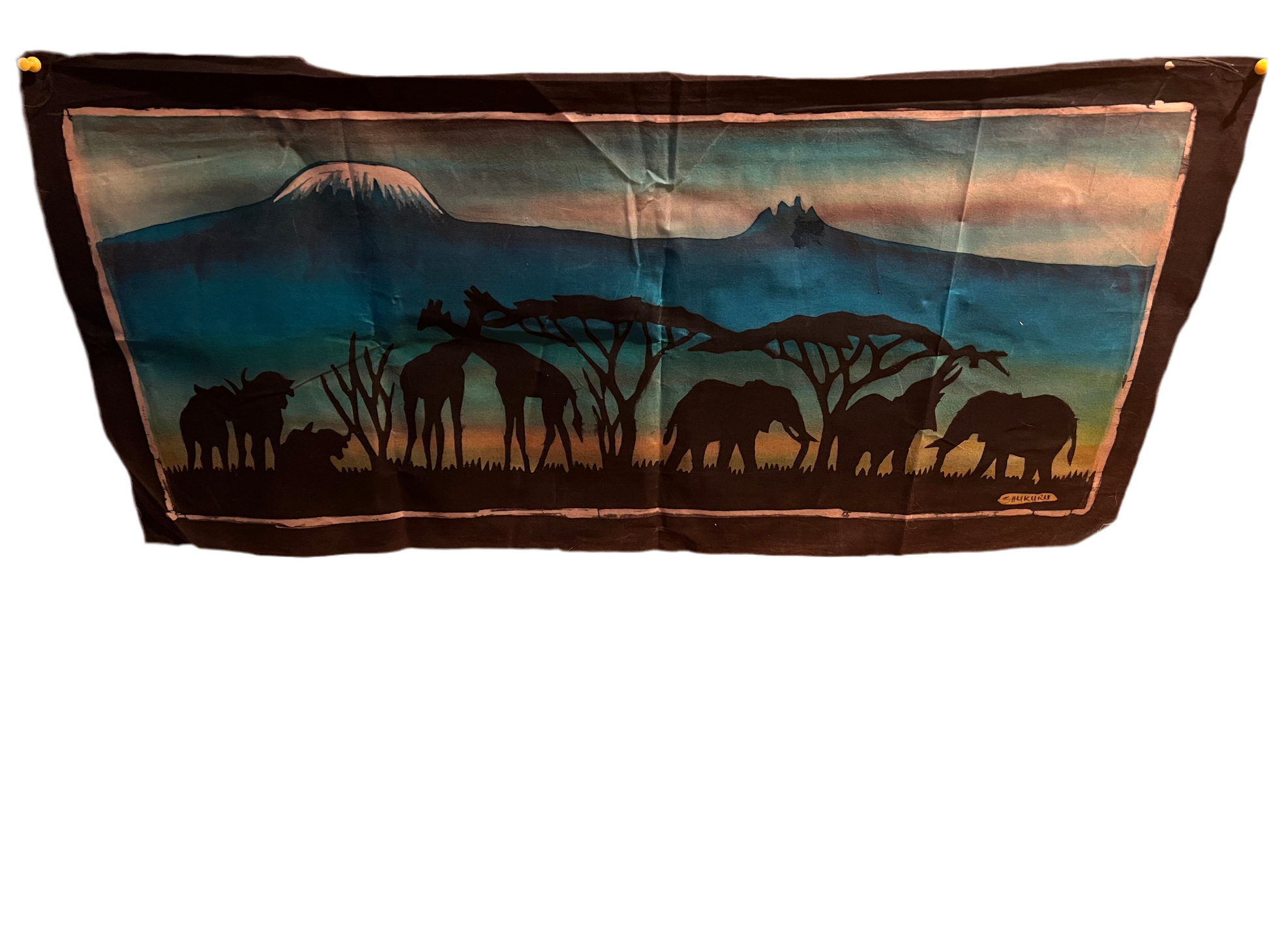 The dynamic artwork "Serengeti Splendor" from Saanskrityam Handicraft Store showcases three abstract figures in vivid attire, each holding a spear against a mountainous background. Surrounding the main piece are various art pieces and masks, skillfully arranged on a textured wall to enhance the overall visual story.