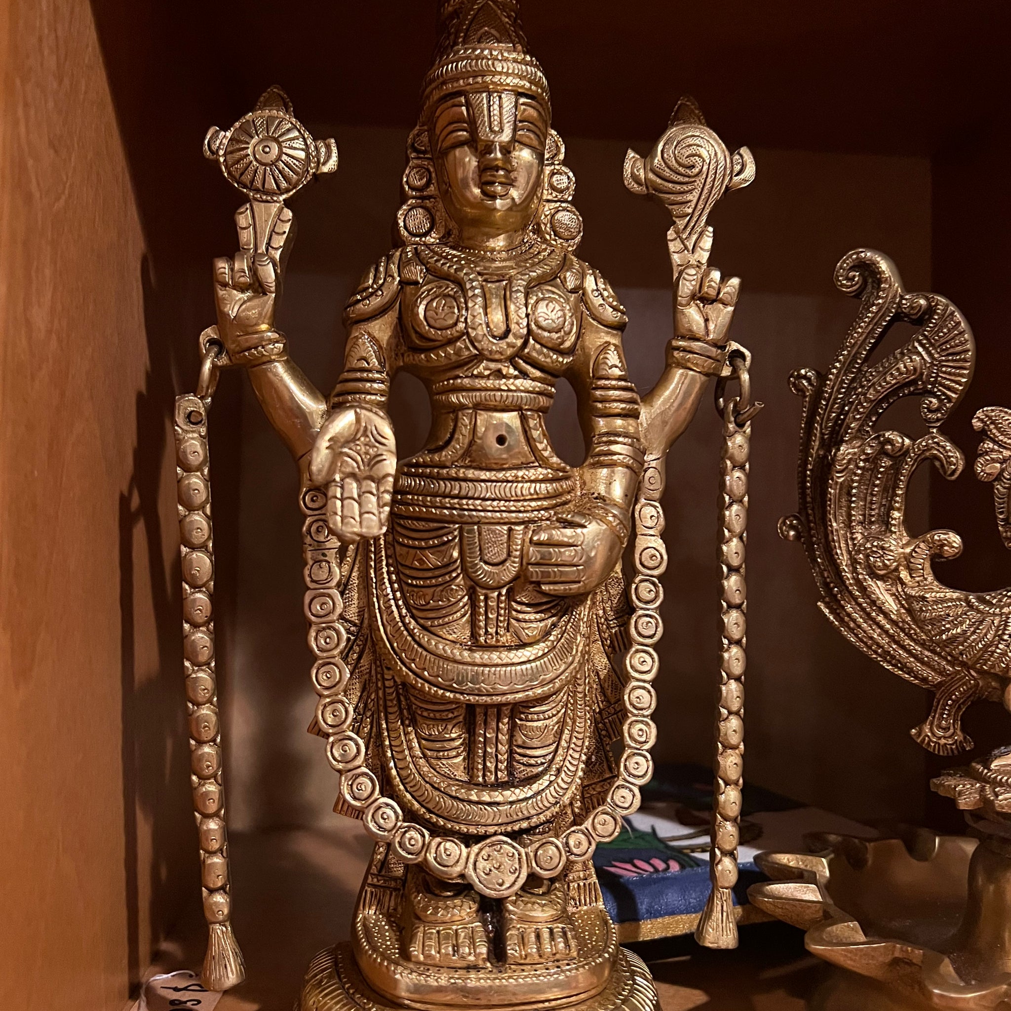 Displayed on a wooden shelf is a bronze Saanskrityam statue from the Balaji Lord of Seven Hills brassware collection, crafted by Saanskrityam Handicraft Store. The figure exudes divine elegance reminiscent of the beauty at Tirumala, holding a lotus in each hand, adorned with intricate jewelry and a tall headdress.