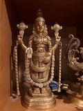 Displayed on a wooden shelf is a bronze Saanskrityam statue from the Balaji Lord of Seven Hills brassware collection, crafted by Saanskrityam Handicraft Store. The figure exudes divine elegance reminiscent of the beauty at Tirumala, holding a lotus in each hand, adorned with intricate jewelry and a tall headdress.