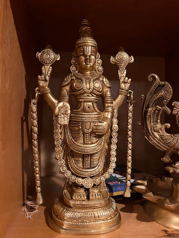 Displayed on a wooden shelf is a bronze Saanskrityam statue from the Balaji Lord of Seven Hills brassware collection, crafted by Saanskrityam Handicraft Store. The figure exudes divine elegance reminiscent of the beauty at Tirumala, holding a lotus in each hand, adorned with intricate jewelry and a tall headdress.