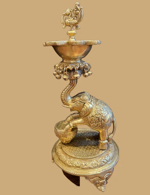 Introducing the Elephant Lamp Brass 18 Inch from Saanskrityam Handicraft Store: A beautifully crafted brass piece showcasing a detailed elephant standing on a circular base. This elegant figure holds a bowl with its raised trunk, adorned by a decorative rooster perched on top.