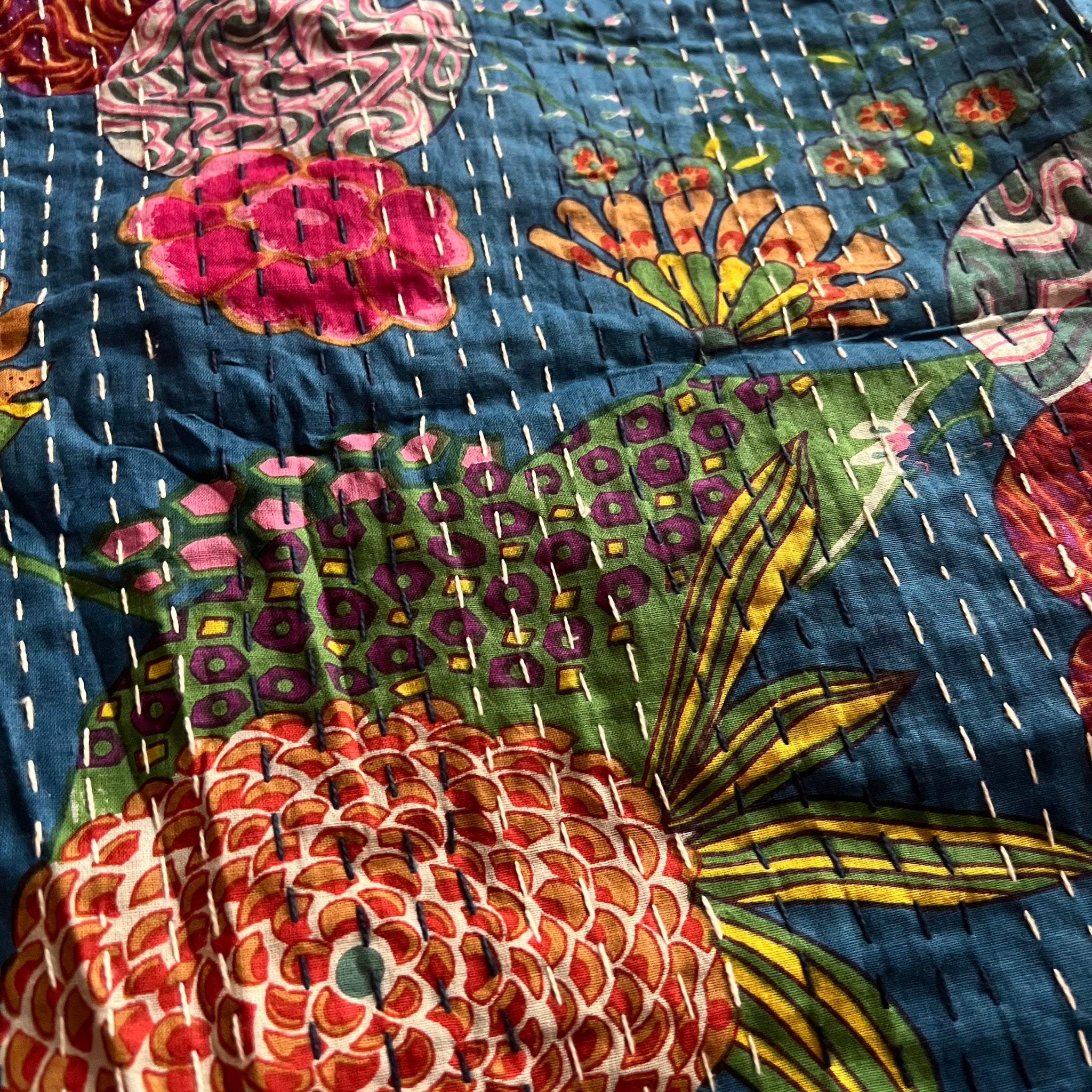 Close-up of the colorful cotton Indian bohemian patchwork quilt from Saanskrityam Handicraft Store, featuring vibrant floral patterns in shades of pink, orange, and green set against a blue background. This king-size design showcases various flowers and leaves with white stitching running vertically across the fabric.