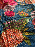 Close-up of the colorful cotton Indian bohemian patchwork quilt from Saanskrityam Handicraft Store, featuring vibrant floral patterns in shades of pink, orange, and green set against a blue background. This king-size design showcases various flowers and leaves with white stitching running vertically across the fabric.