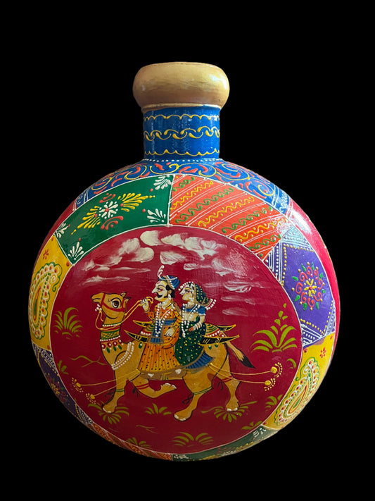 Experience the vibrant beauty of Saanskrityam Handicraft Store's "Rajasthani Art on Metal Vase." This handcrafted piece from India features colorful traditional artwork, with a central image depicting a man and woman in traditional attire riding a camel and intricate patterns in various bright colors surrounding them.