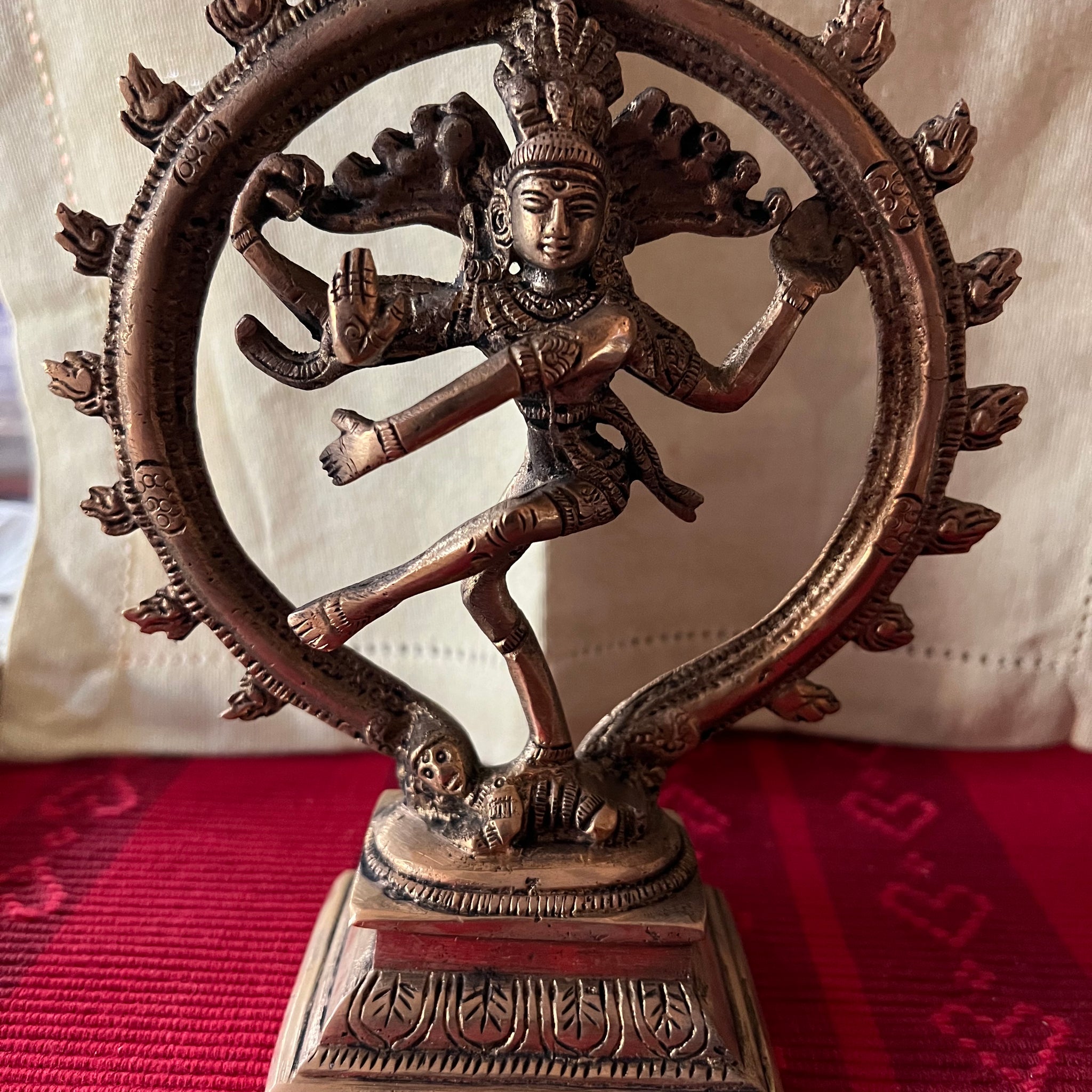 Shiva statue from the Tamil Nadu brassware Shivoham collection, part of Saanskrityam Handicraft Store, featuring the Hindu deity in a cosmic dance pose encircled by a ring of flames. This intricately detailed brass figure rests on a red and gold patterned cloth background.