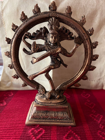 Shiva statue from the Tamil Nadu brassware Shivoham collection, part of Saanskrityam Handicraft Store, featuring the Hindu deity in a cosmic dance pose encircled by a ring of flames. This intricately detailed brass figure rests on a red and gold patterned cloth background.