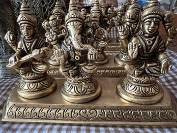 A set of intricately carved brass statues from the Ganesha, Lakshmi Saraswathi collection by Saanskrityam Handicraft Store is arranged on a checkered cloth. These statues feature detailed embellishments and are seated on a rectangular base, beautifully depicting Hindu deities.