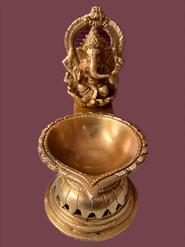 The Ganesh lamp 7 inch from Saanskrityam Handicraft Store is a brass oil lamp showcasing an intricate design with the deity Ganesha gracefully seated above a circular bowl adorned with ornate edges. It boasts a traditional, antique appearance and is beautifully set against a maroon background.