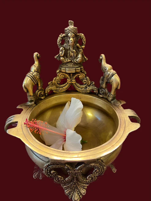 A 7-inch brass Ganesh urli from Saanskrityam Handicraft Store, featuring intricate carvings of a deity accompanied by two elephants, holding a white hibiscus flower against a maroon background.