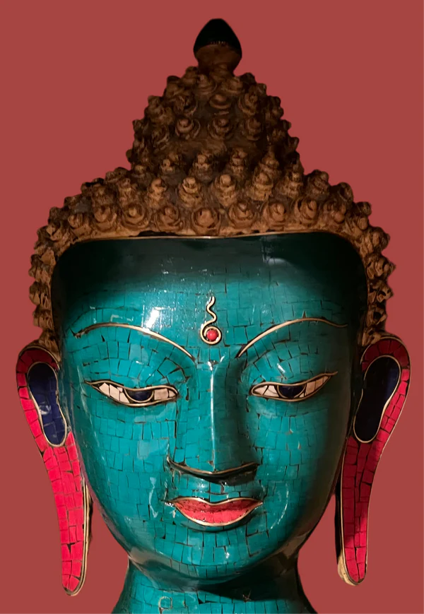 A vibrant green lapis Buddha head mask, crafted by Saanskrityam Handicraft Store, features a serene expression with curly hair and is enhanced with red and blue accents on the ears, red lips, and a striking red jewel on the forehead against a solid red background.