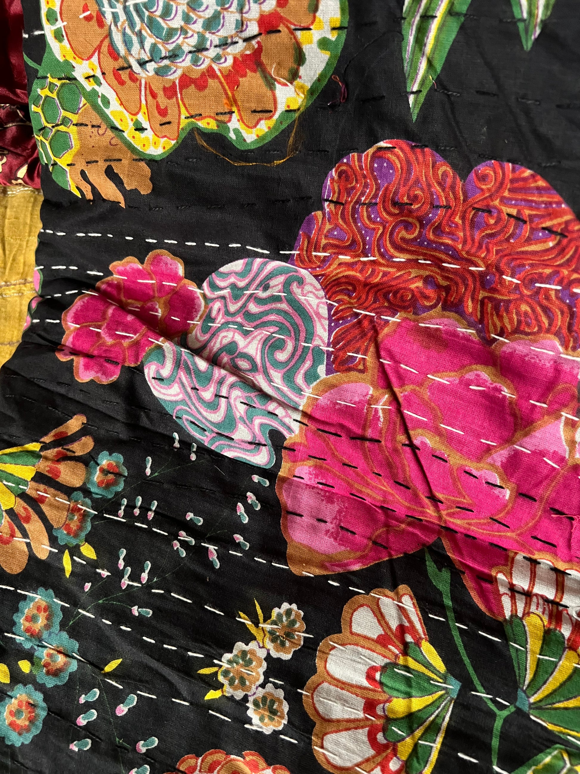 Close-up of the colorful cotton Indian bohemian patchwork quilt from Saanskrityam Handicraft Store, featuring vibrant floral patterns in shades of pink, orange, and green set against a blue background. This king-size design showcases various flowers and leaves with white stitching running vertically across the fabric.