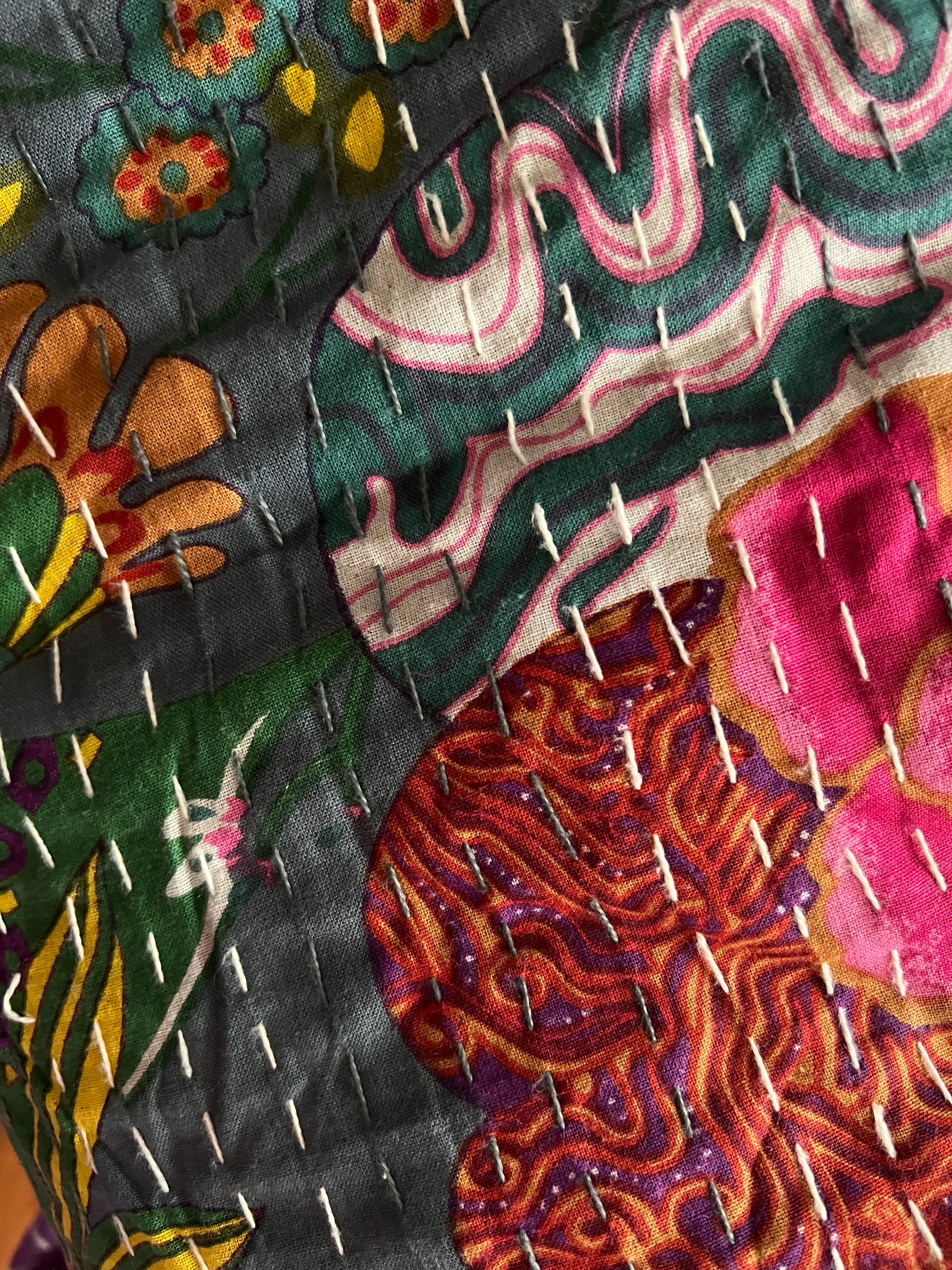 Close-up of the colorful cotton Indian bohemian patchwork quilt from Saanskrityam Handicraft Store, featuring vibrant floral patterns in shades of pink, orange, and green set against a blue background. This king-size design showcases various flowers and leaves with white stitching running vertically across the fabric.