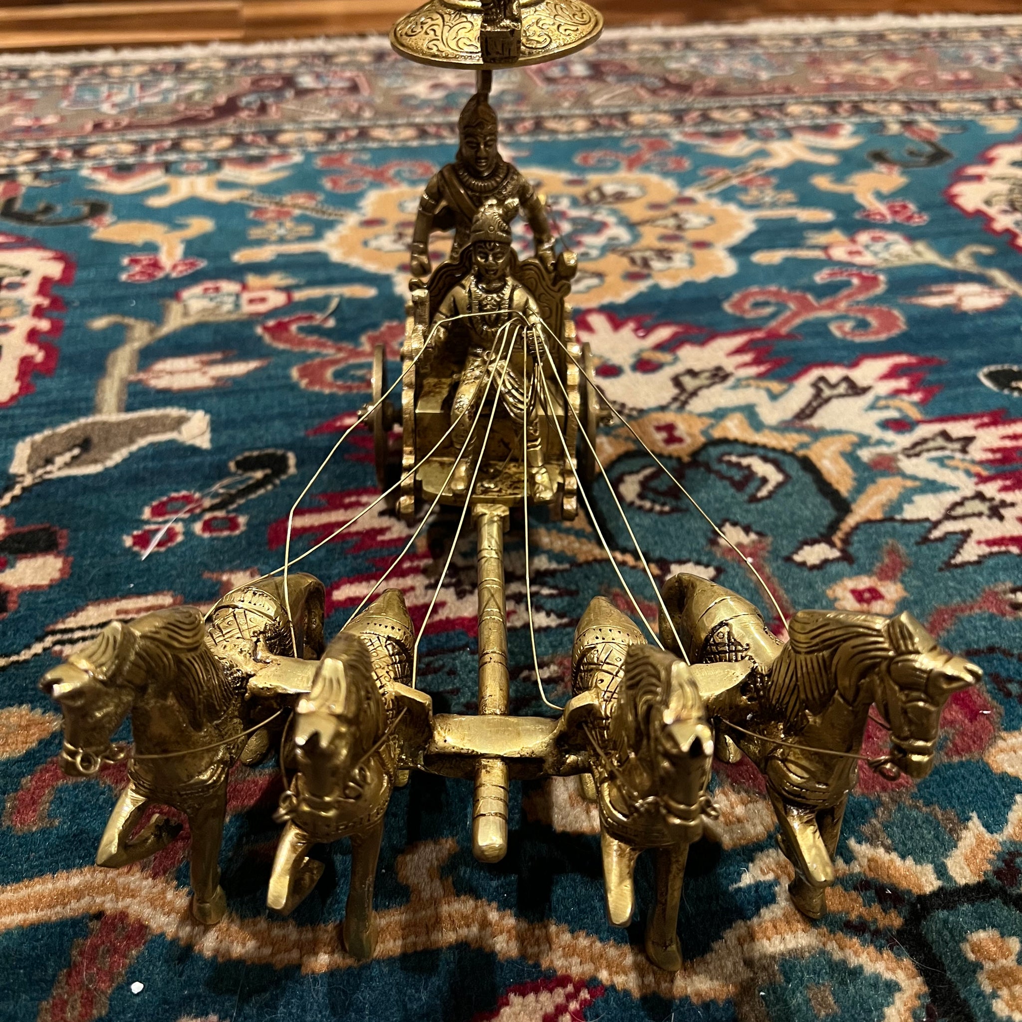 A bronze chariot sculpture named "Krishna and Arjuna / Bhagavad Gita at Kurukshetra," crafted by the Saanskrityam Handicraft Store, depicting four horses with Arjuna and Krishna holding a bow under an umbrella, is elegantly placed on an intricately patterned blue and red rug.