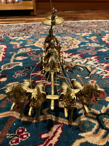 A bronze chariot sculpture named "Krishna and Arjuna / Bhagavad Gita at Kurukshetra," crafted by the Saanskrityam Handicraft Store, depicting four horses with Arjuna and Krishna holding a bow under an umbrella, is elegantly placed on an intricately patterned blue and red rug.