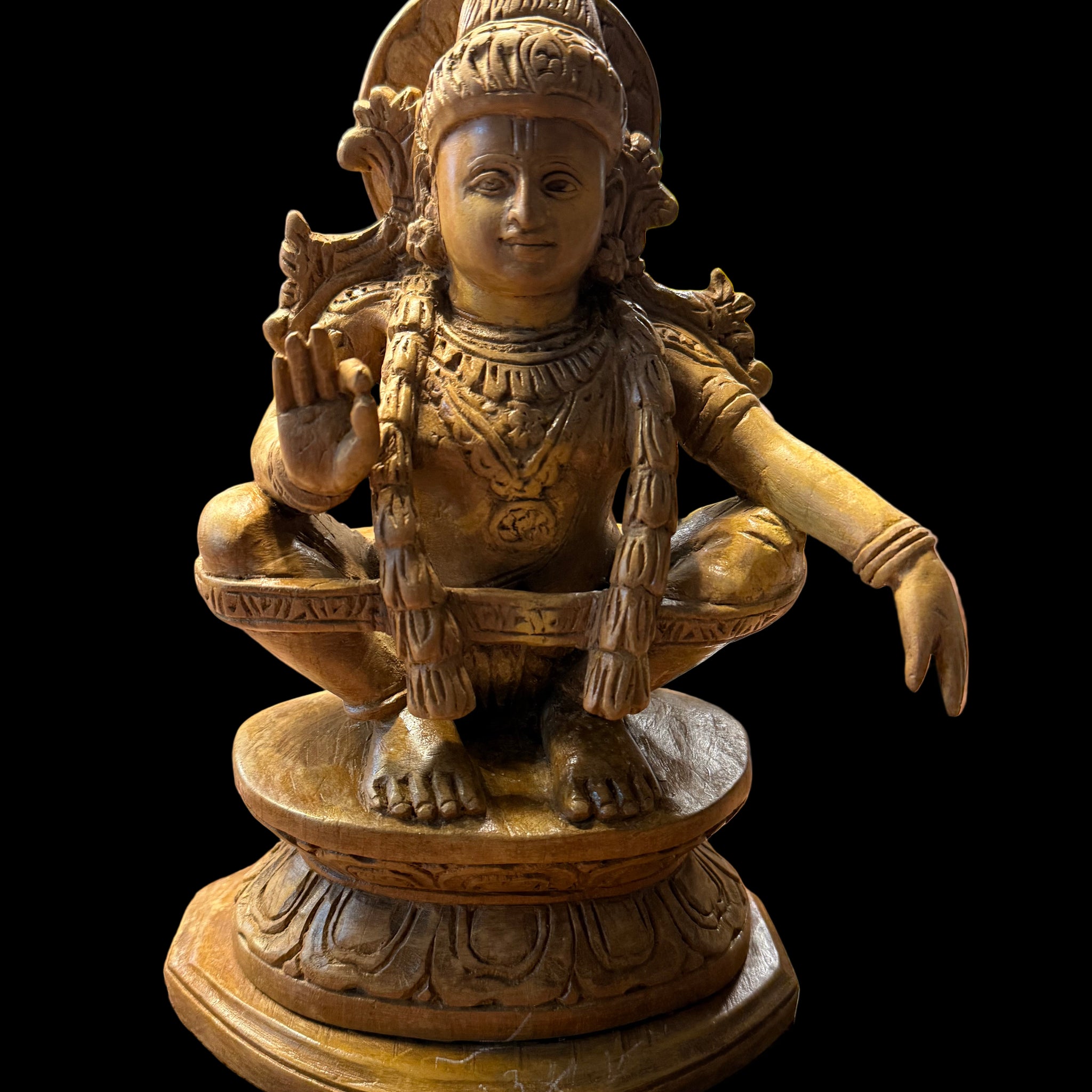A 7-inch "Sri Ayyappa bronze with arch" statue from Saanskrityam Handicraft Store is displayed on a wooden shelf. The deity is depicted in a meditative pose, enclosed within an ornate bronze arch, with intricate details adorning both the figure and base. Other decorative objects can be partially seen in the background.