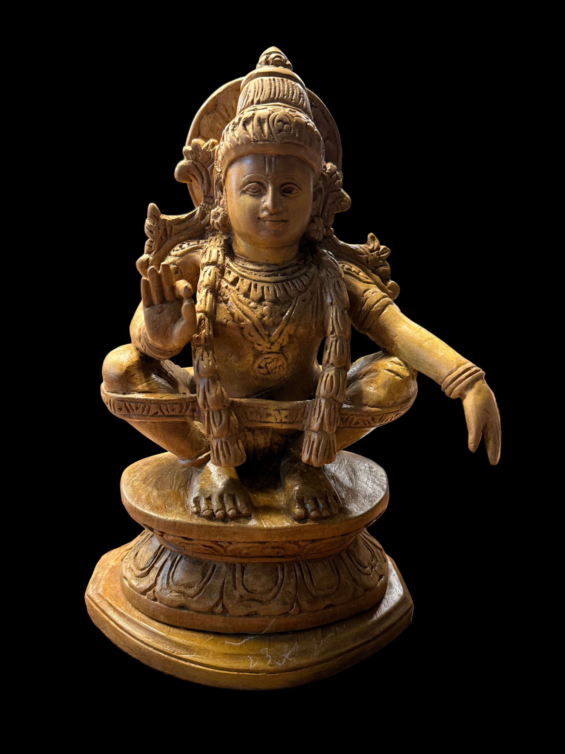 A 7-inch "Sri Ayyappa bronze with arch" statue from Saanskrityam Handicraft Store is displayed on a wooden shelf. The deity is depicted in a meditative pose, enclosed within an ornate bronze arch, with intricate details adorning both the figure and base. Other decorative objects can be partially seen in the background.