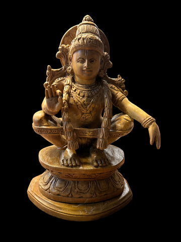 A 7-inch "Sri Ayyappa bronze with arch" statue from Saanskrityam Handicraft Store is displayed on a wooden shelf. The deity is depicted in a meditative pose, enclosed within an ornate bronze arch, with intricate details adorning both the figure and base. Other decorative objects can be partially seen in the background.