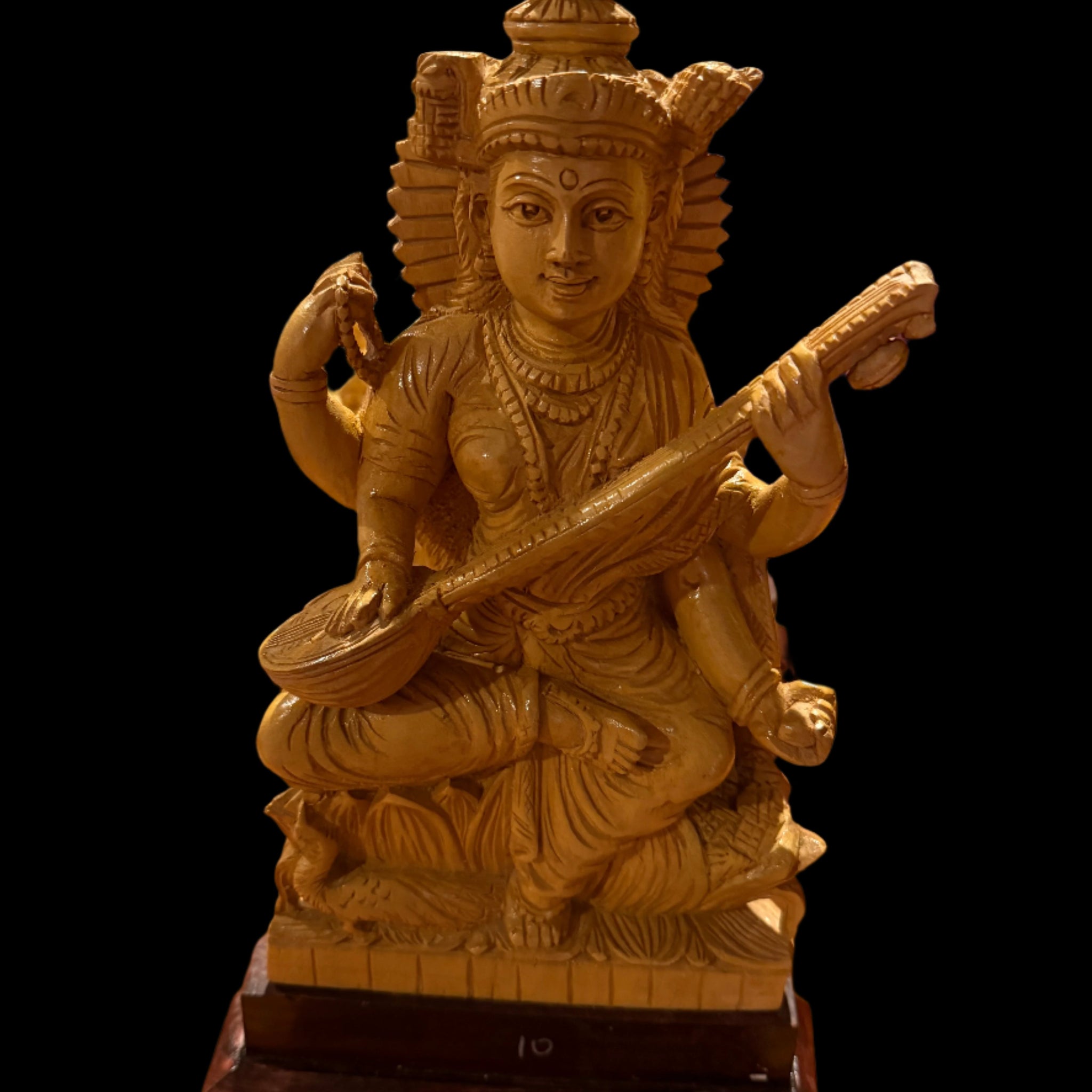 A handcrafted wood Saraswathi statue, measuring 12 inches and offered by Saanskrityam Handicraft Store, showcases the deity with multiple arms, playing a stringed instrument. The statue features intricately detailed attire and a headdress, depicted gracefully seated on a small pedestal.