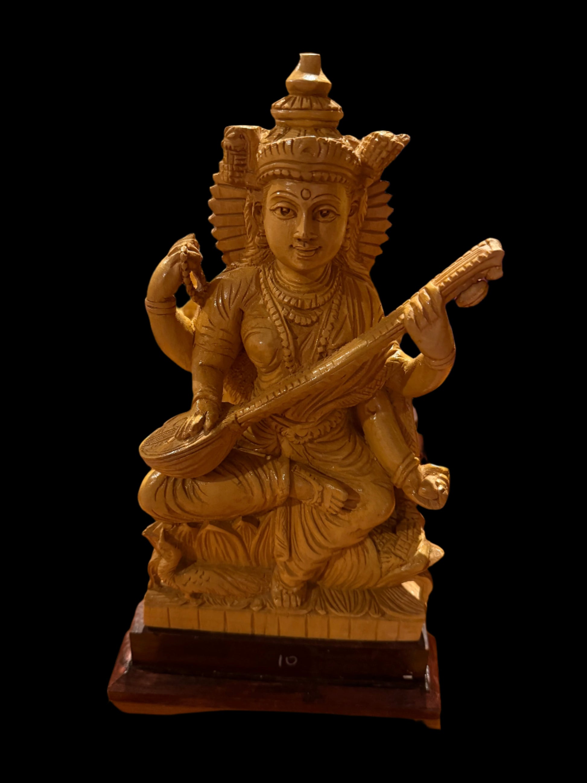 A handcrafted wood Saraswathi statue, measuring 12 inches and offered by Saanskrityam Handicraft Store, showcases the deity with multiple arms, playing a stringed instrument. The statue features intricately detailed attire and a headdress, depicted gracefully seated on a small pedestal.