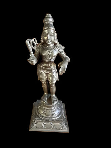 A bronze statue of Krishna by Saanskrityam Handicraft Store stands on an ornate base. The figure features intricate carvings, with a raised hand and traditional attire, all set against a plain black background.