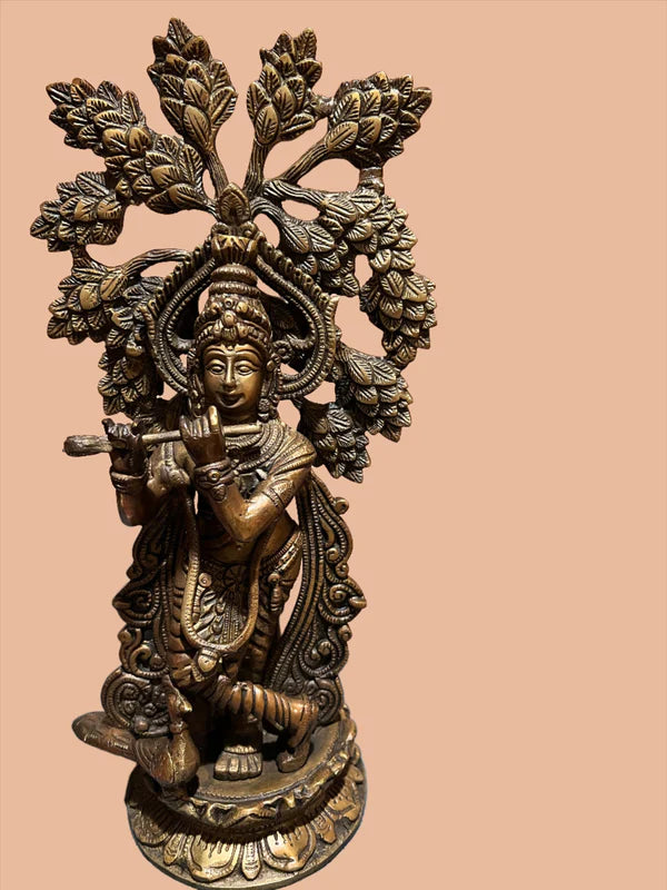 Product Description: Introducing the Krishna bronze 12 inch statue by Saanskrityam Handicraft Store. This exquisite piece captures a serene figure playing a flute, standing gracefully beneath an intricately carved tree. The figure is adorned in ornate clothing and features a peaceful expression, set against a solid, soft peach background.