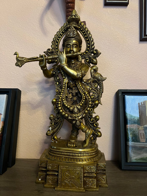 The Krishna bronze 20-inch statue from Saanskrityam Handicraft Store depicts a Hindu deity playing a flute. It is beautifully adorned with intricate patterns and stands elegantly on a decorative pedestal. The figure showcases traditional attire along with ornate details and is positioned against a pale wall flanked by framed paintings.