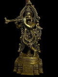 The Krishna bronze 20-inch statue from Saanskrityam Handicraft Store depicts a Hindu deity playing a flute. It is beautifully adorned with intricate patterns and stands elegantly on a decorative pedestal. The figure showcases traditional attire along with ornate details and is positioned against a pale wall flanked by framed paintings.