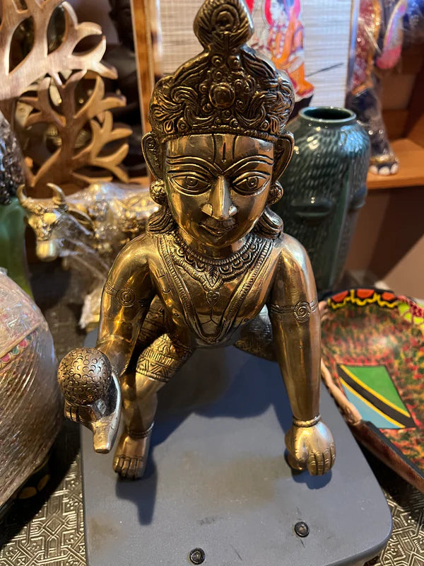 A bronze statue named "Laddoo Gopal" crafted by Saanskrityam Handicraft Store, measuring 14 inches, elegantly depicts a deity with an ornate headdress and garments, poised on one knee while holding a spherical object. This intricate piece is accentuated by decorative items like vases, a wooden animal figure, and a colorful tray.