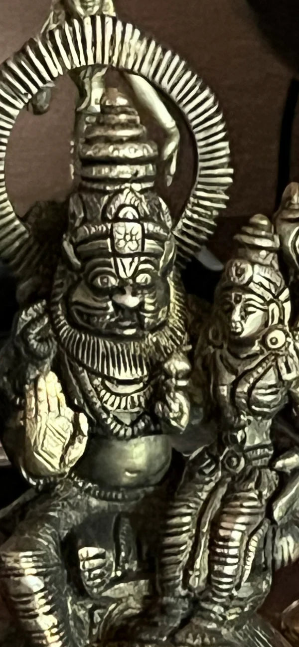 The Narasimhaswamy with Lakshmi Devi statue from Saanskrityam Handicraft Store is a 6-inch brass piece with copper hues, showcasing a detailed scene from Hindu mythology. The central deity, with multiple arms and a lion-like face, is seated next to another figure adorned with elaborate headdresses and ornamentation.