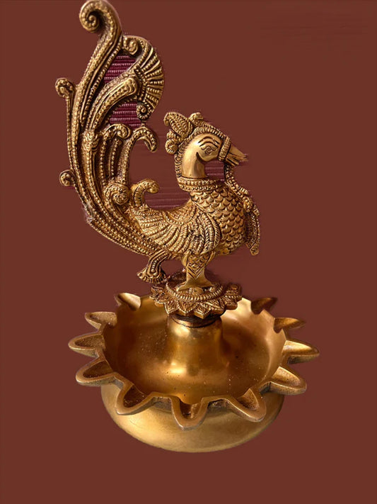 A brass peacock lamp from the Saanskrityam Handicraft Store, measuring 9 inches, showcases an intricately carved peacock design. The detailed feathers of the peacock stand atop a shallow dish with a sunburst pattern on the edge, all set against a plain brown background.