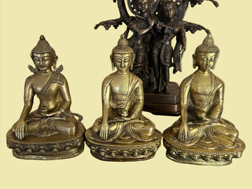 The Set of 3 Buddha bronzes from Saanskrityam Handicraft Store features three meditative statues, each seated on decorative bases. They are positioned in front of a richly detailed, multi-armed deity in a dark hue, contrasted by a gentle yellow backdrop.