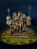 A Shiv Parivaar heavy bronze sculpture from Saanskrityam Handicraft Store, depicting the deity riding a bull and holding a trident, is placed on a beautifully decorated plate adorned with intricate leaf patterns. Against a dark blue backdrop, two wooden objects and a metallic trident complete the scene.