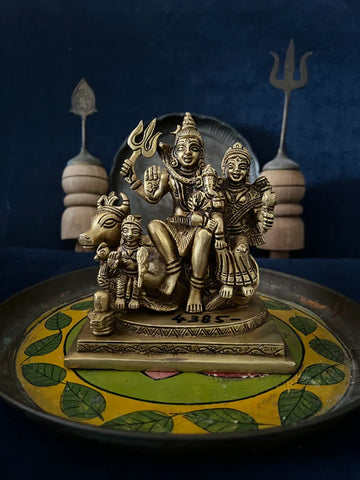 A Shiv Parivaar heavy bronze sculpture from Saanskrityam Handicraft Store, depicting the deity riding a bull and holding a trident, is placed on a beautifully decorated plate adorned with intricate leaf patterns. Against a dark blue backdrop, two wooden objects and a metallic trident complete the scene.