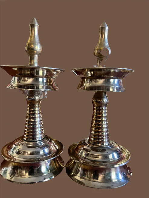 Displayed against a plain brown background are two small-sized simple Kerala standing lamps from Saanskrityam Handicraft Store. These intricately designed brass lamps feature conical bases and pointed tops, with a polished, reflective surface that highlights their detailed craftsmanship.