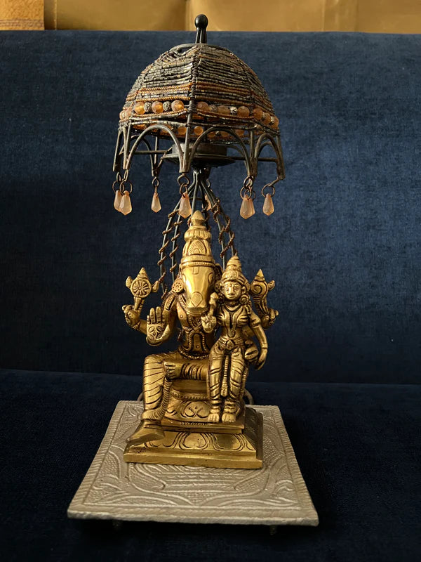 The Sri Varahaswamy with Lakshmi Devi 6-inch bronze statue from Saanskrityam Handicraft Store gracefully showcases two seated deities beneath a beautifully crafted umbrella adorned with intricate patterns. The base is embellished with embossed designs, contributing an elegant touch to the overall artistry.
