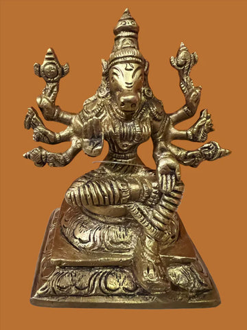 The Varaahi Devi 6-inch bronze statue by Saanskrityam Handicraft Store portrays a multi-armed deity seated with an intricately detailed costume and headdress, resting on a decorative pedestal against a warm orange background.