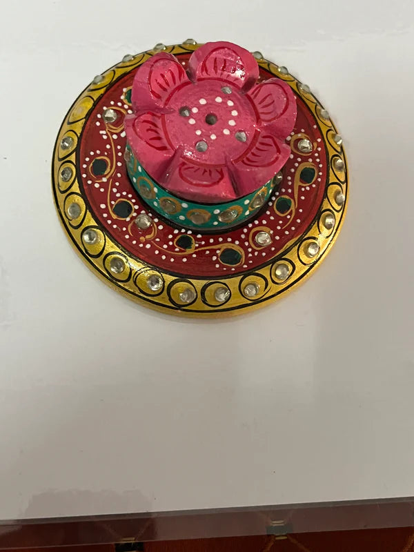 The "Marble Agarbatti Stand Colorful" from Saanskrityam Handicraft Store features a decorative round base with intricate yellow and red patterns, adorned with small reflective stones. Atop the base is a pink lotus-shaped ornament accented with green, also embellished with small white stones, all placed elegantly on a white surface.