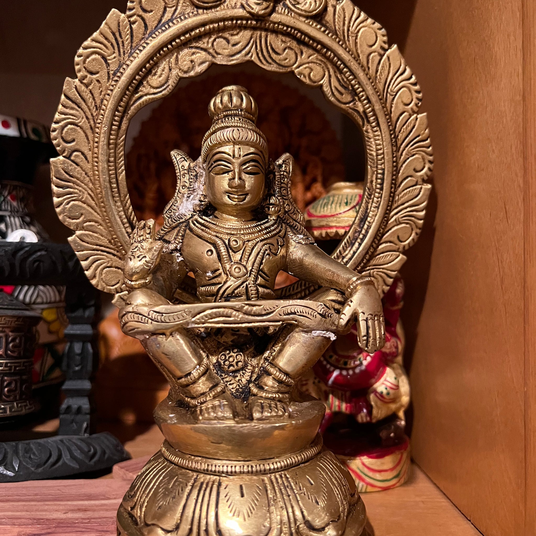 A 7-inch "Sri Ayyappa bronze with arch" statue from Saanskrityam Handicraft Store is displayed on a wooden shelf. The deity is depicted in a meditative pose, enclosed within an ornate bronze arch, with intricate details adorning both the figure and base. Other decorative objects can be partially seen in the background.