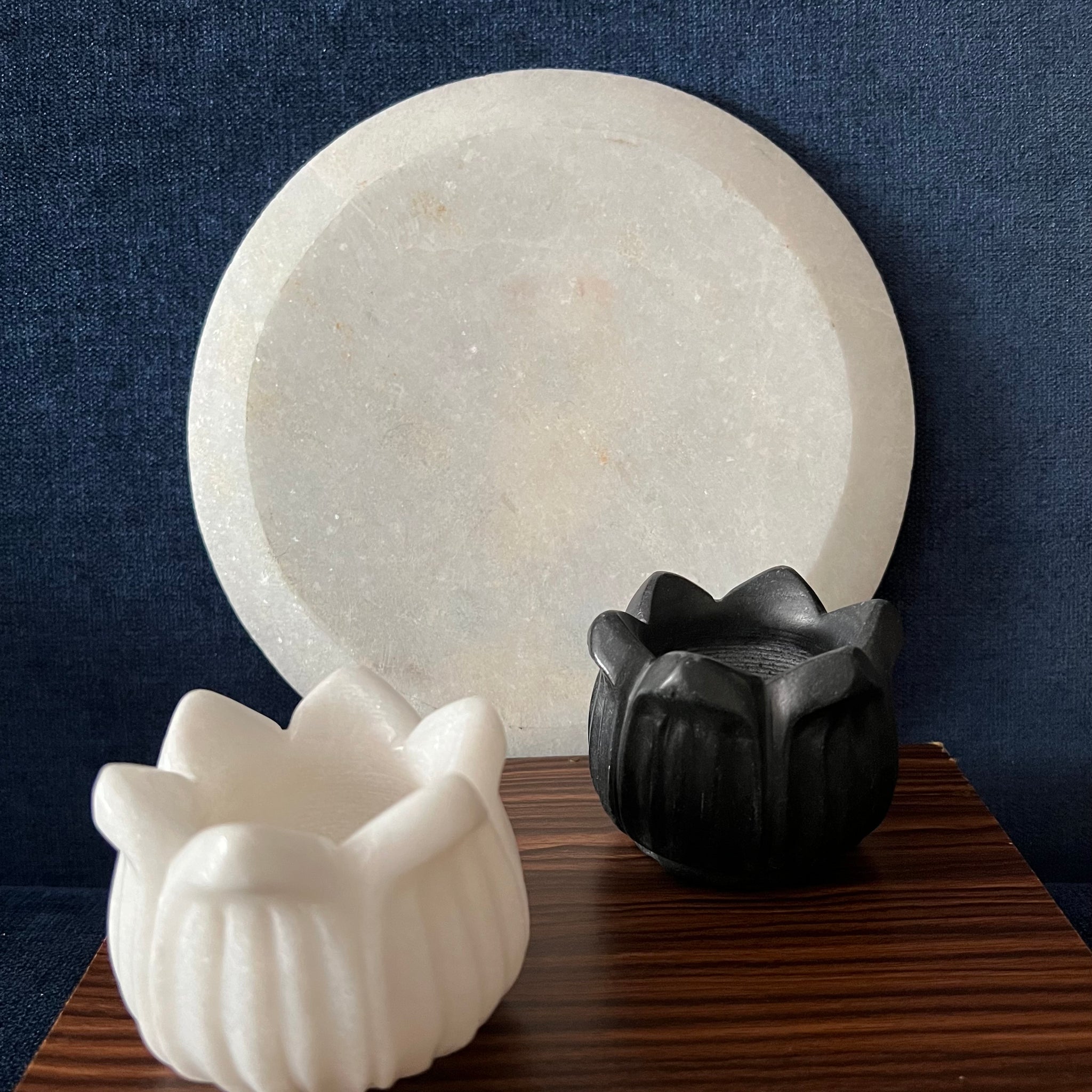 A black and white flower-shaped agarbatti stand is positioned in front of a round candle holder from the Saanskrityam Handicraft Store's Marble Candle Stands collection. Both holders rest on a wooden surface set against a dark blue background.