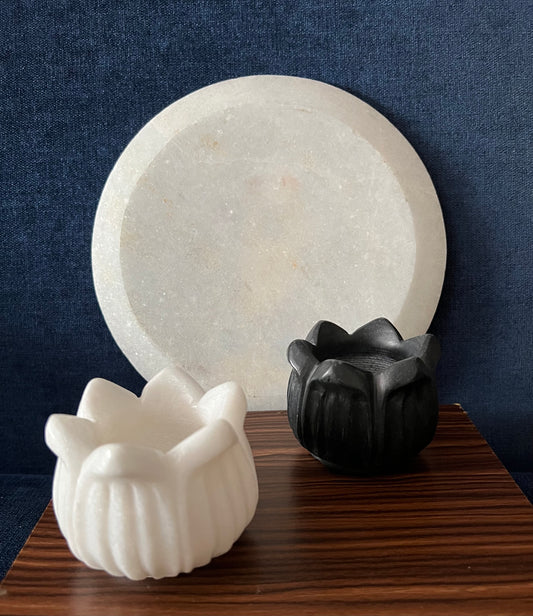 A black and white flower-shaped agarbatti stand is positioned in front of a round candle holder from the Saanskrityam Handicraft Store's Marble Candle Stands collection. Both holders rest on a wooden surface set against a dark blue background.