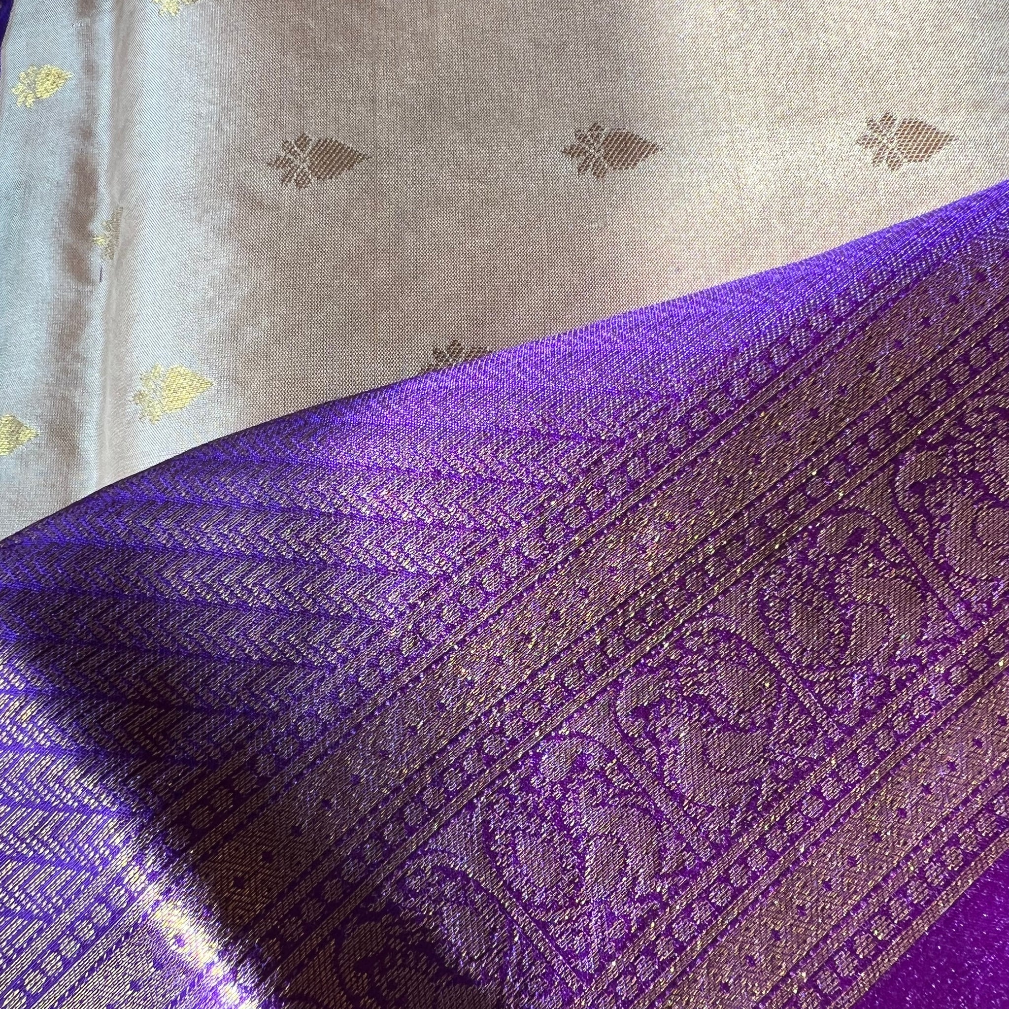 A close-up of the luxurious Kanchipuram silk saree from Saanskrityam Handicraft Store showcases an elegant purple section with intricate gold patterns, paired with a silver section adorned with delicate gold motifs. The pure silk fabric reflects light beautifully, emphasizing its exquisite texture and detailed design typical of high-quality silk sarees.