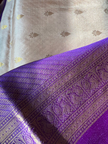 A close-up of the luxurious Kanchipuram silk saree from Saanskrityam Handicraft Store showcases an elegant purple section with intricate gold patterns, paired with a silver section adorned with delicate gold motifs. The pure silk fabric reflects light beautifully, emphasizing its exquisite texture and detailed design typical of high-quality silk sarees.