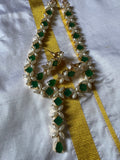 A piece from Saanskrityam Handicraft Store's collection, the Jewellery/Indian cosmetic jewellery with stones and metal, featuring a gold chain entwined with vibrant green gemstones reminiscent of traditional Indian style, is beautifully draped over a textured wooden surface. The muted dark background enhances the contrast and richness of this exquisite piece.