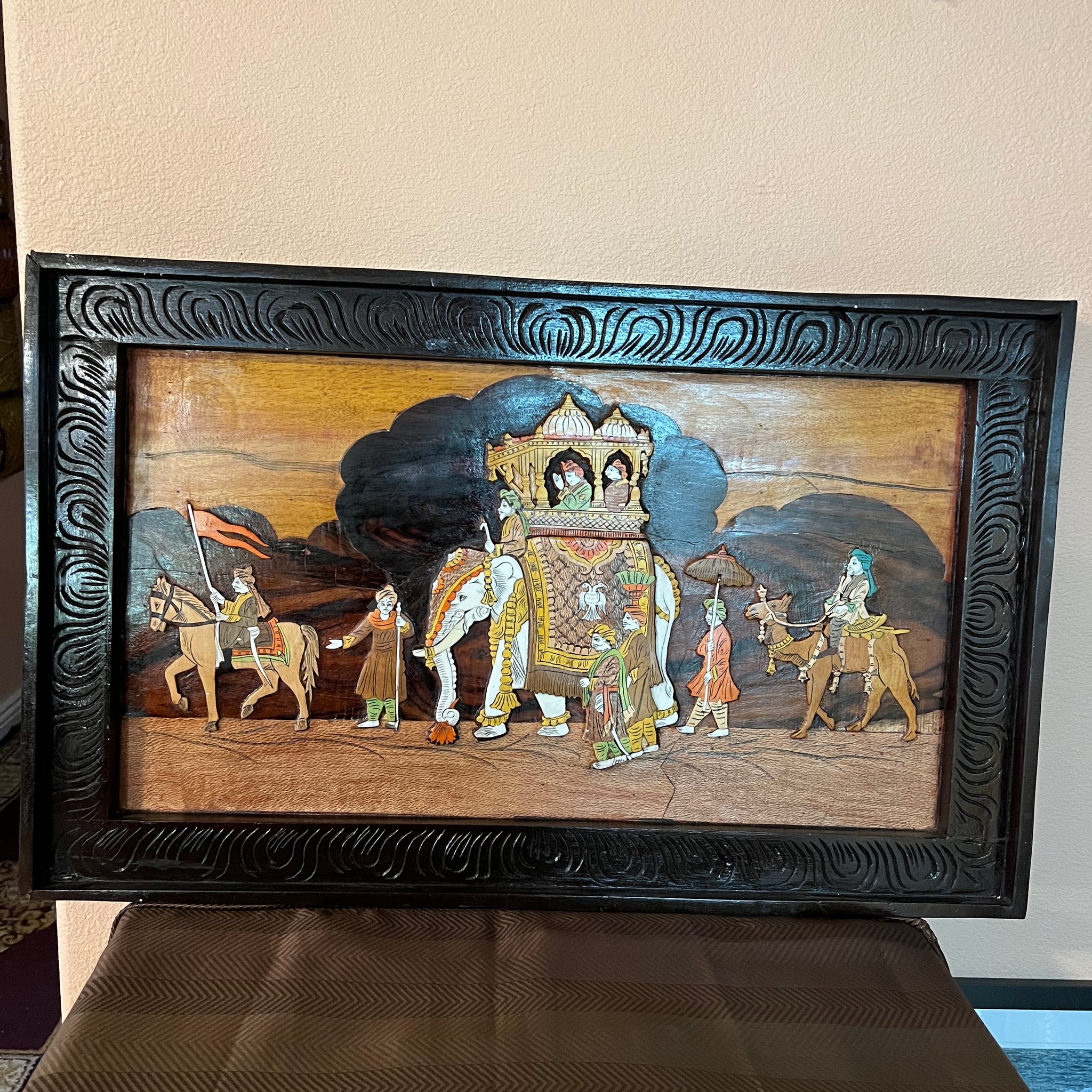 A framed piece of Karnataka rosewood art from the Saanskrityam Handicraft Store depicts a traditional royal procession. The scene includes a chariot with two seated figures, an elephant guided by attendants, and people on foot against a stylized backdrop. The ornate frame is intricately carved from rosewood.