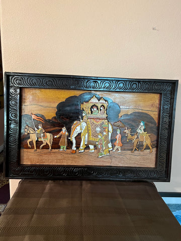 A framed piece of Karnataka rosewood art from the Saanskrityam Handicraft Store depicts a traditional royal procession. The scene includes a chariot with two seated figures, an elephant guided by attendants, and people on foot against a stylized backdrop. The ornate frame is intricately carved from rosewood.