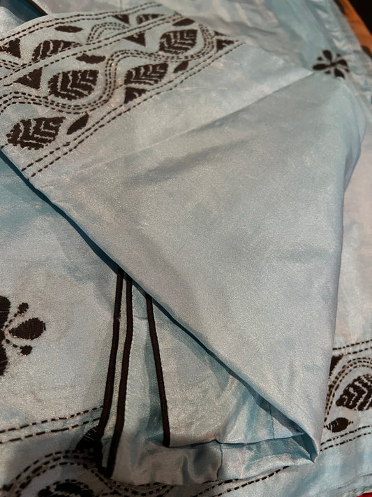 Detailed view of the Kantha thread work Bangla silk saree from Saanskrityam Handicraft Store, featuring intricate black threadwork that forms elegant floral and vine patterns on a light blue background. The shiny texture enhances its appeal, with a folded section at the top. This fabric includes a ready-to-wear blouse, making it an excellent choice for sophisticated attire.