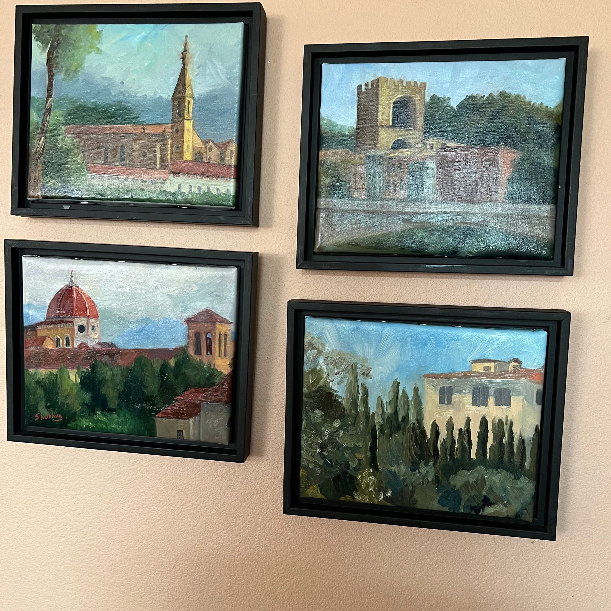 Four framed paintings adorn the wall, each showcasing distinct architectural scenes featuring churches and buildings amidst trees and blue skies. The vibrant colors and expressive brushwork create a scenic, picturesque atmosphere reminiscent of Shubhra Sarkar's style. These pieces belong to "Saanskrityam" by contemporary Indian artist Shubhra Sarkar from the Saanskrityam Handicraft Store, beautifully capturing the essence of Florence.