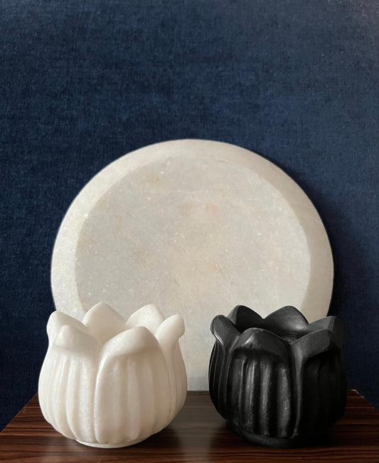 A black and white flower-shaped agarbatti stand is positioned in front of a round candle holder from the Saanskrityam Handicraft Store's Marble Candle Stands collection. Both holders rest on a wooden surface set against a dark blue background.